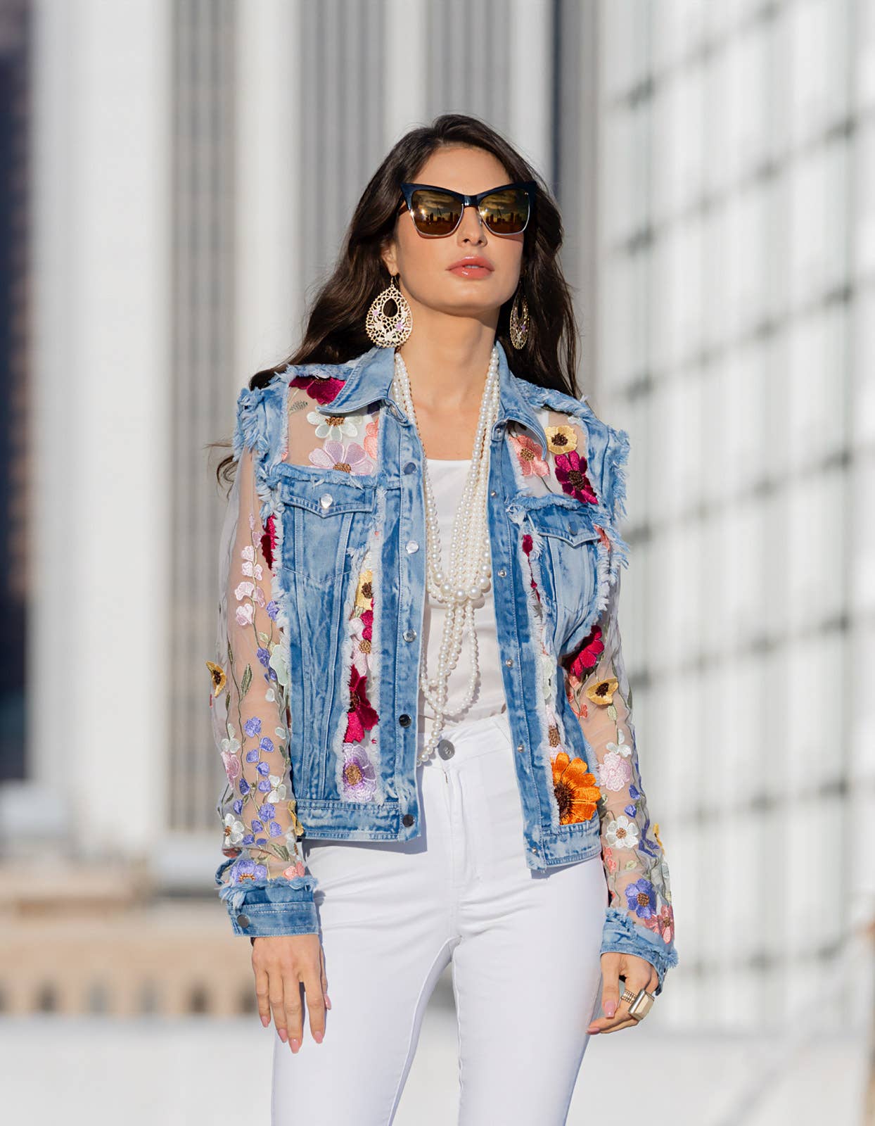 Woman in sunglasses wearing floral denim jacket and white pants.