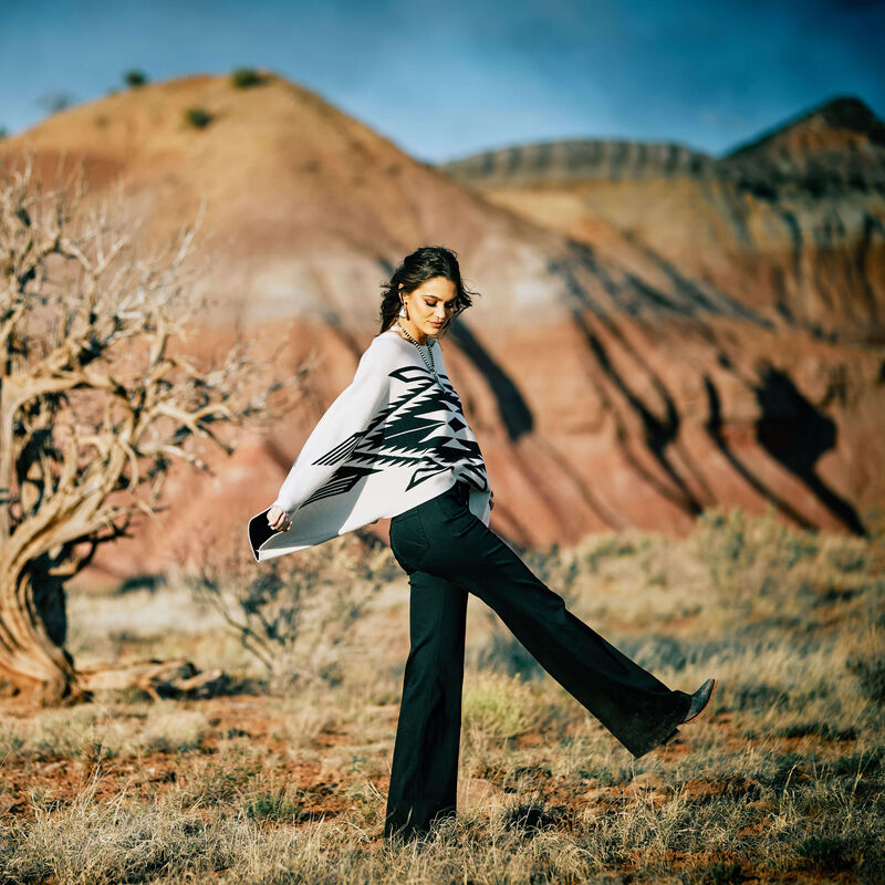 Woman in patterned top and black pants, with red mountains.