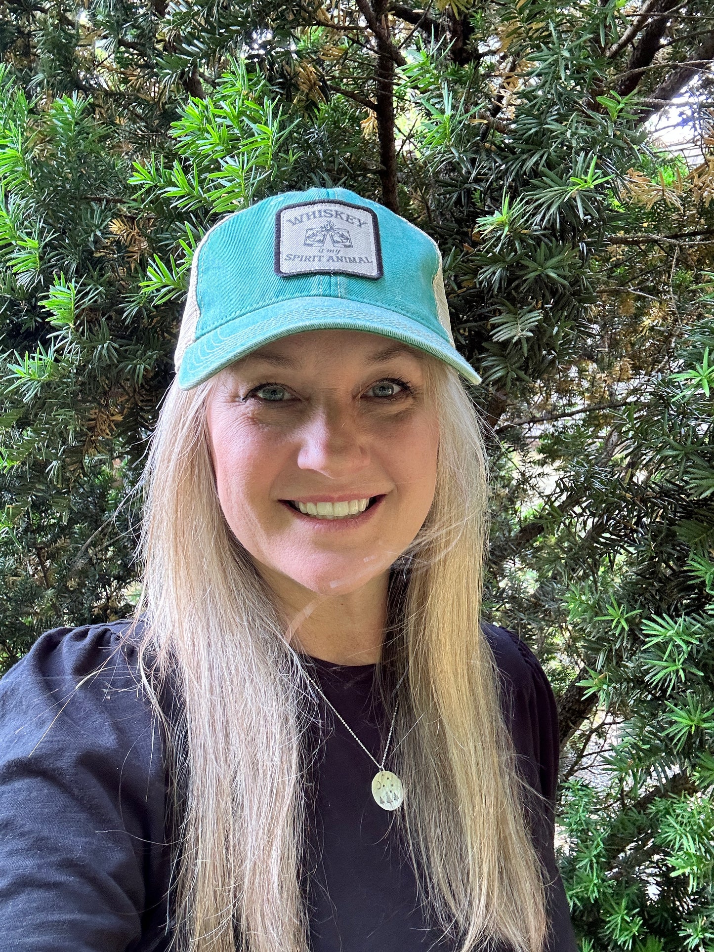Woman smiling, wearing a teal hat with a patch, outdoors.