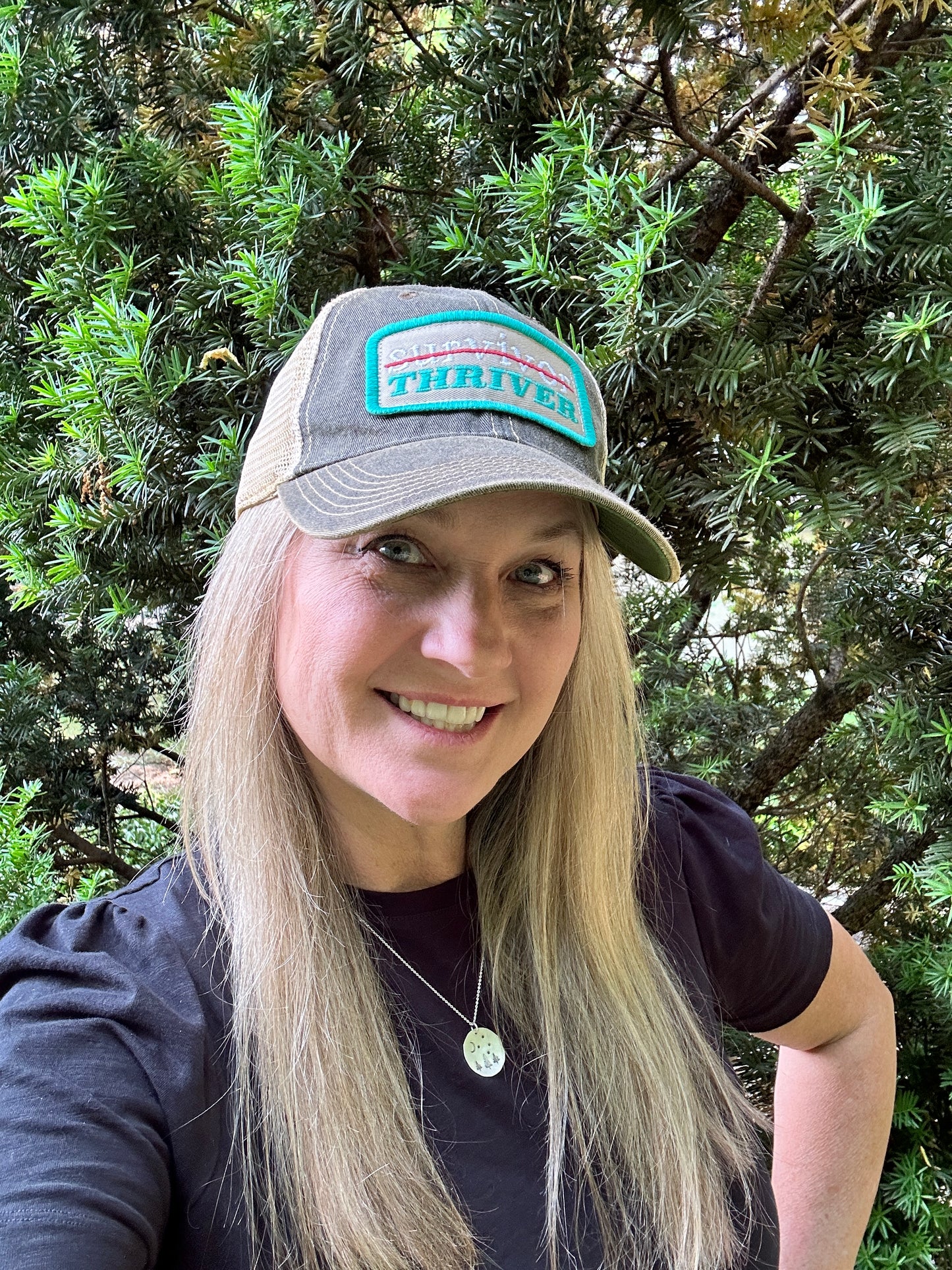 Woman wearing a hat and smiling in front of greenery.