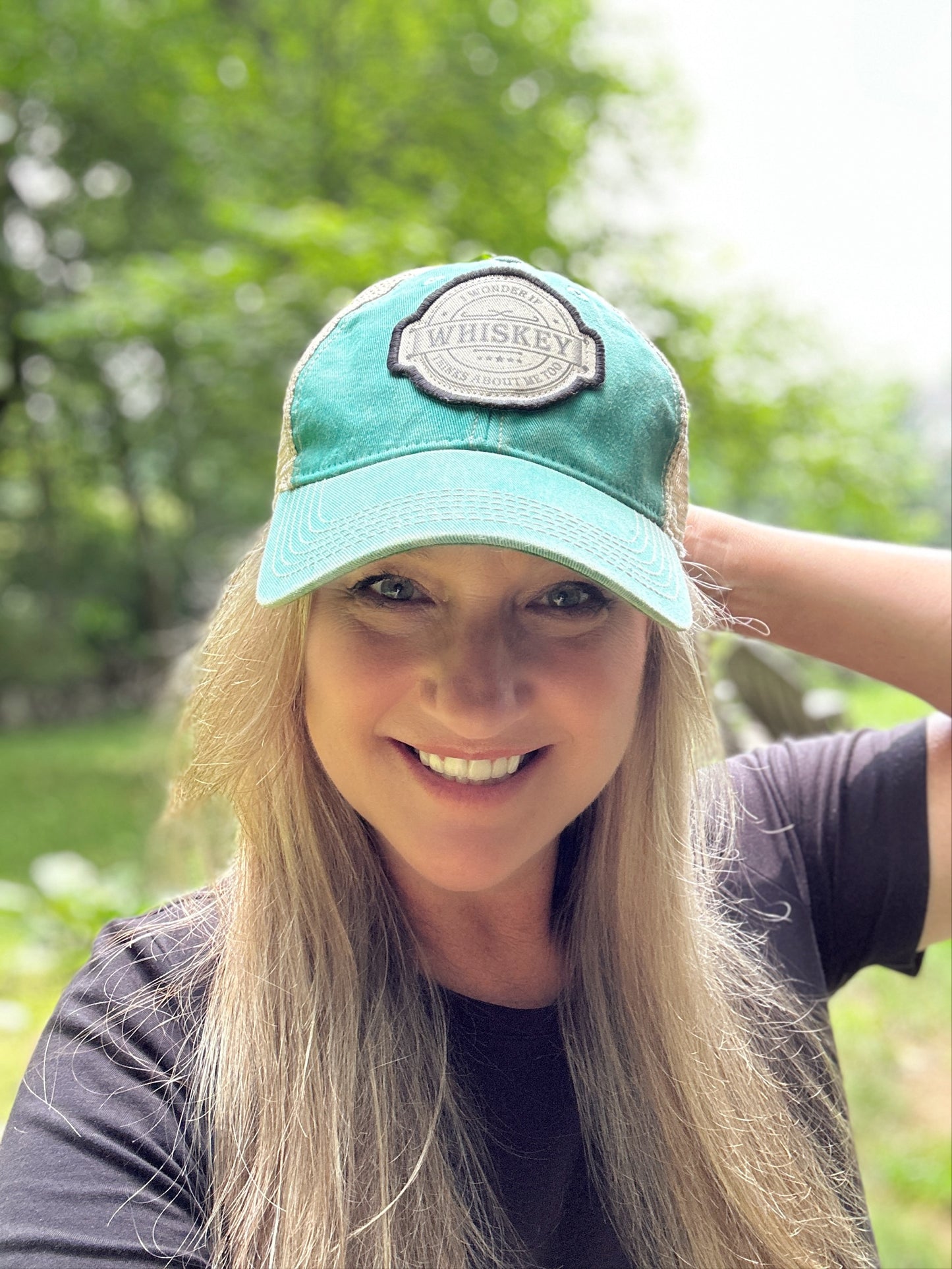Woman smiling, wearing teal hat with Whiskey patch.