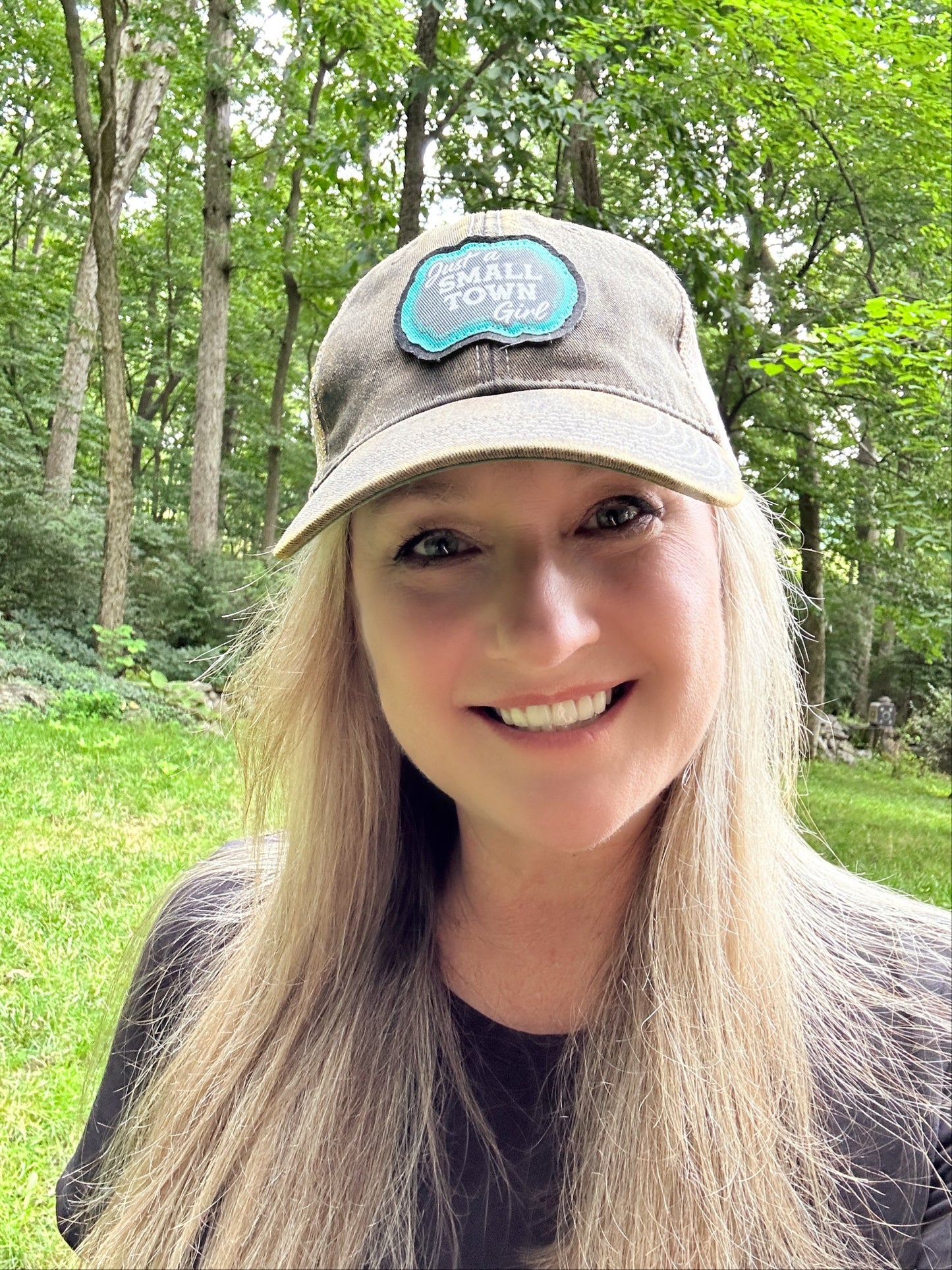 Woman smiling, wearing a Small Town Good cap in a forest.