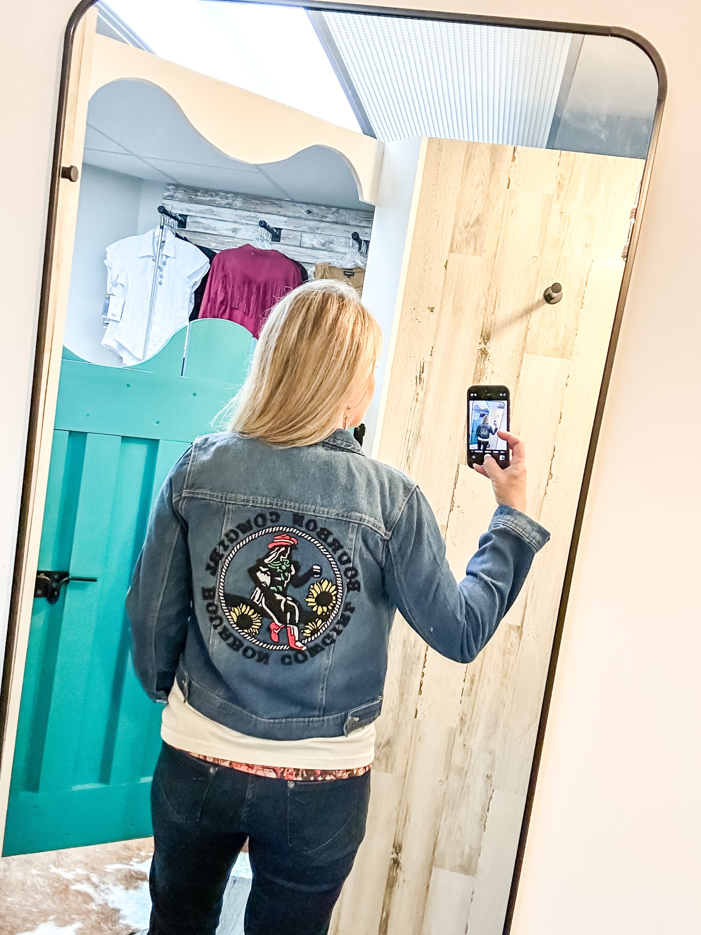 Woman in denim jacket taking a photo in mirror.