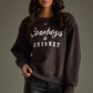 Woman wearing a brown Cowboys & Whiskey sweatshirt and black pants.